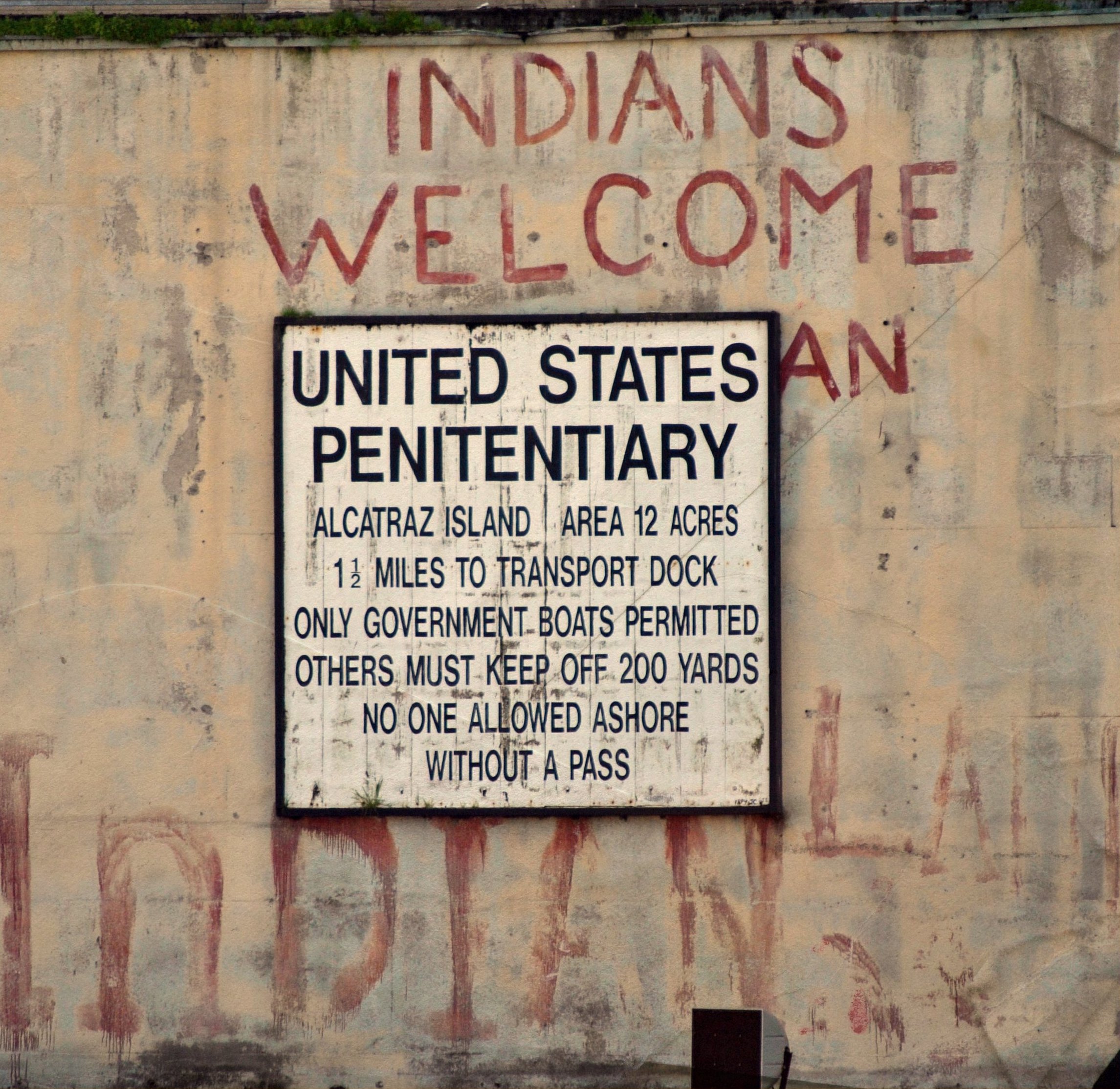 historical photo shows graffiti, "Indians welcome Indians," on a sign that says "Federal Penitentiary, Alcatraz Island, Area 12 Acres, 1.5 miles to transport dock, only government boats permitted, others must keep off 200 yards, no one allowed ashore without a pass."