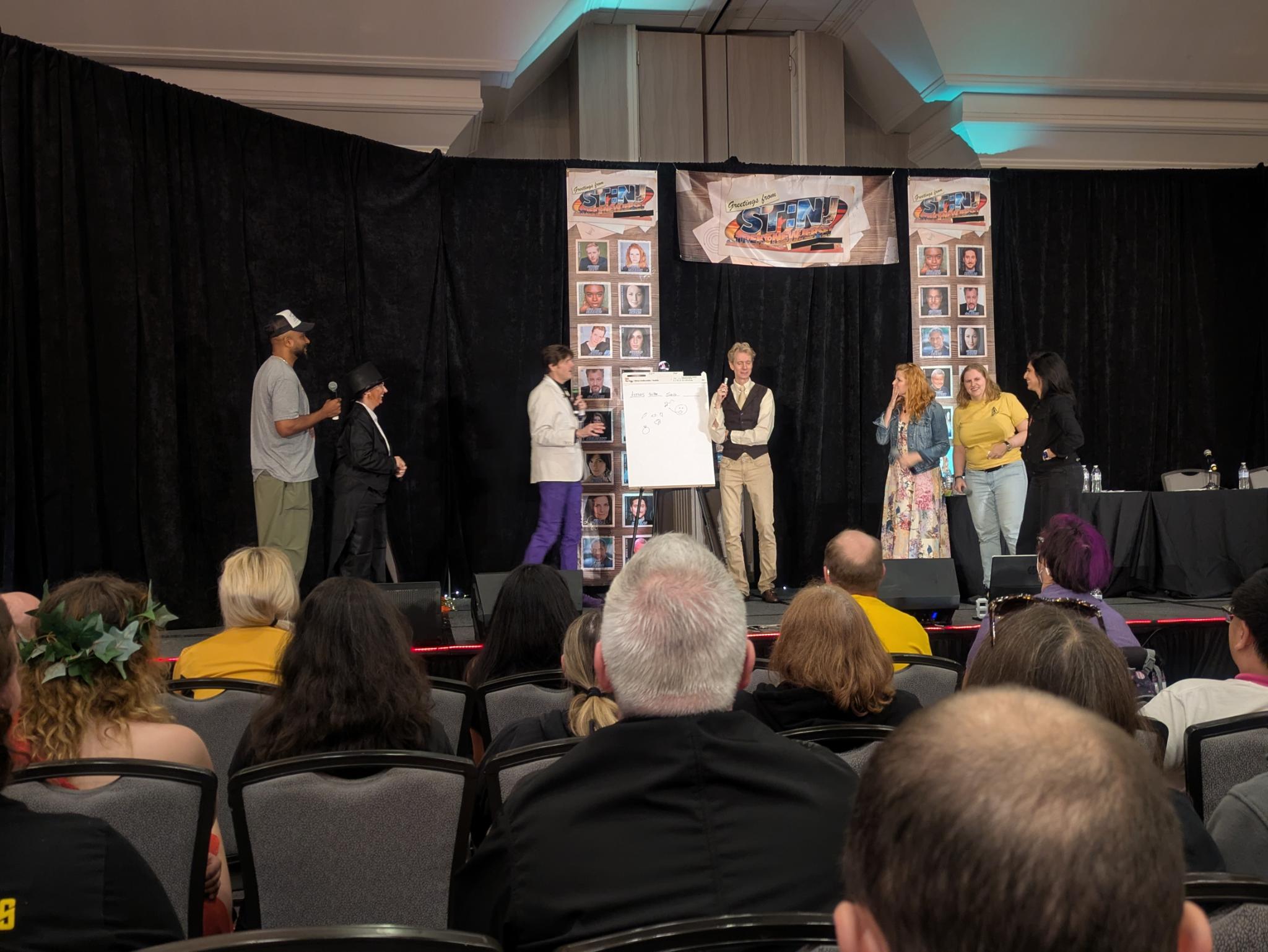 A stage with Cirroc Lofton, a fan dressed as Abraham Lincoln, the MC, a pictionary board, Doug Jones drawing on it, and Bonnie Gordon, me, and Gabrielle Ruiz