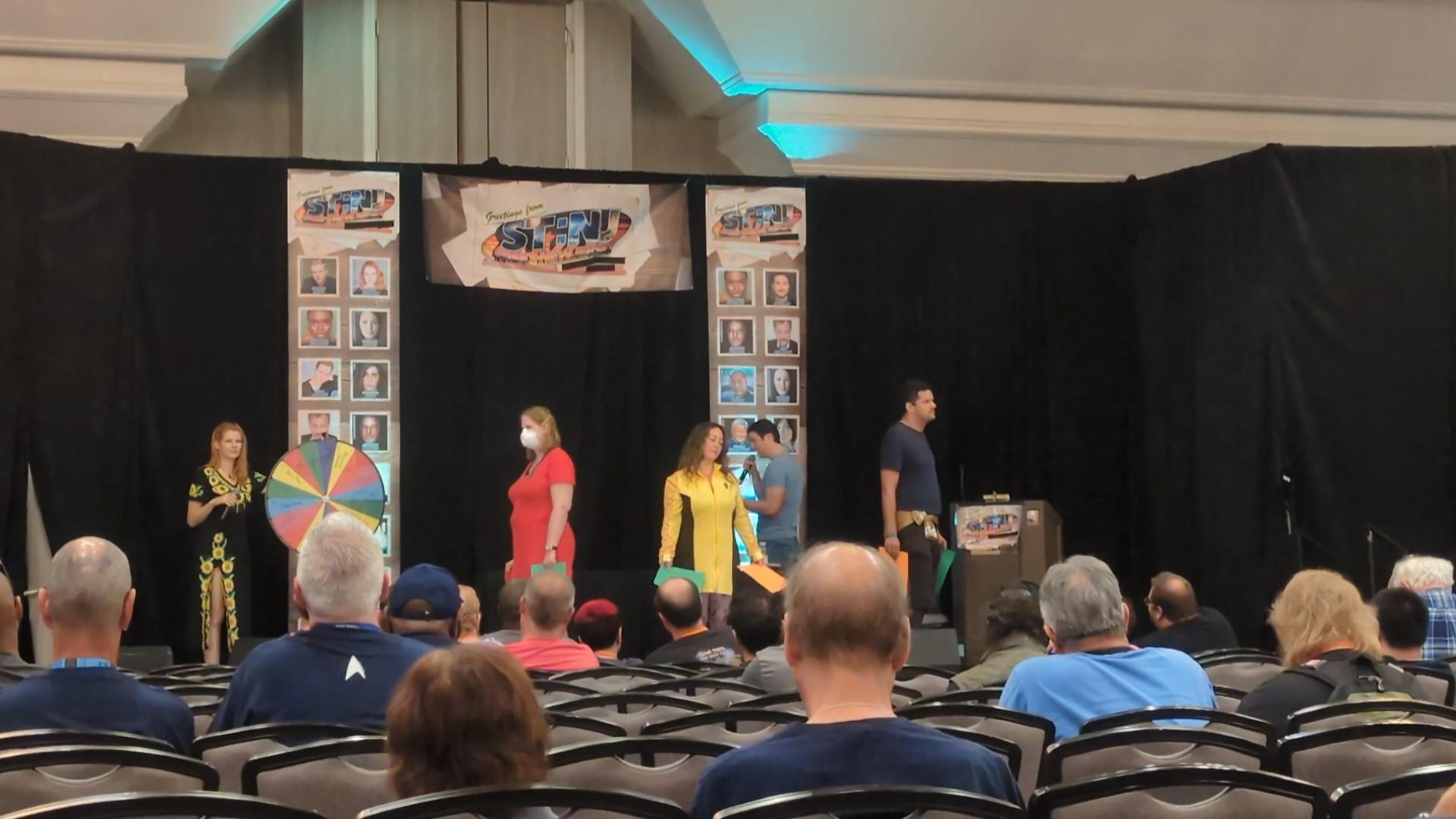 picture description: a stage with a colorful wheel, Bonnie Gordon in a sunflower dress, and me, a woman in a command gold dress, a man with a microphone, and a man not in a costume