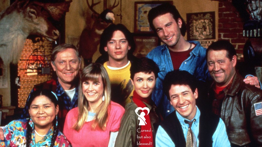 Picture description: group photo of 8 characters from Northern Exposure. Maggie's marked with our logo and the words "cursed - but also blessed?"