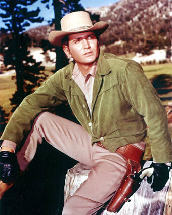 A white man wearing a light brown cowboy hat, green long-sleeved jacket, black gloves, left-handed leather holster, and tan pants.