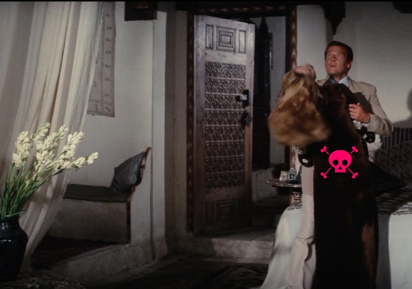 Roger Moore stares up in shock while the woman he's embracing slumps backwards. The site's pink-skull logo is on her back.