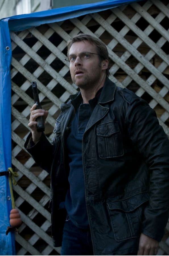 Michael Shanks in-character, holding a gun and wearing a leather jacket over jeans and a t-shirt