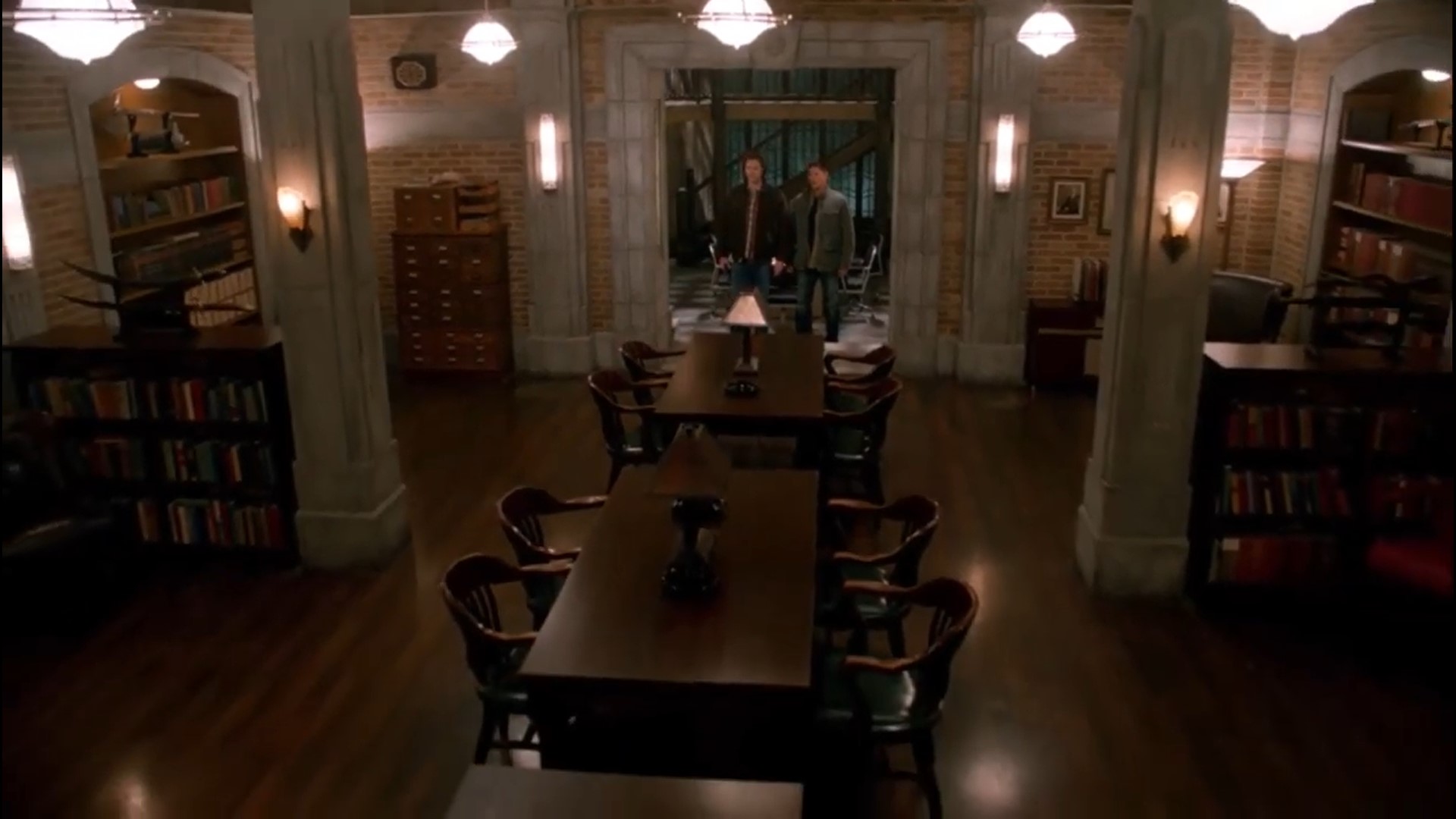 Sam and Dean staring in wonder at a huge room full of stuffed bookshelves, with a long conference table down the middle